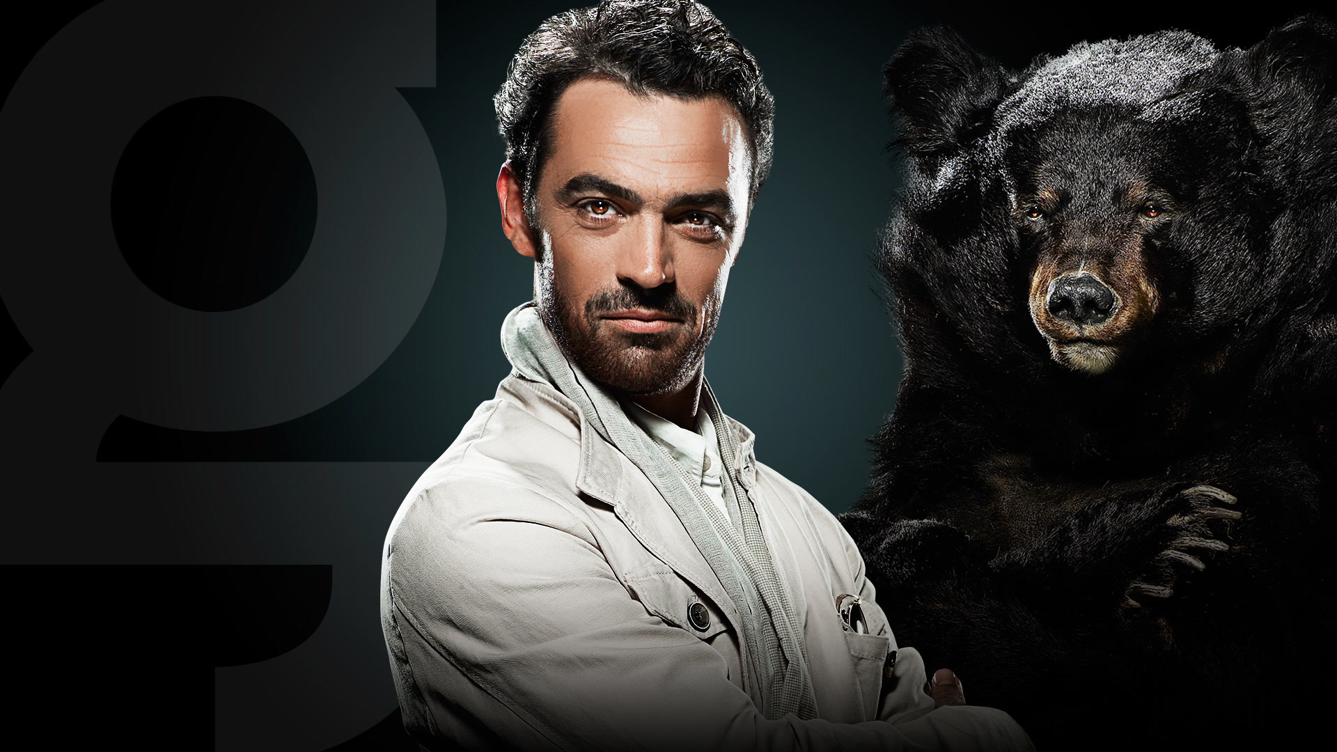 Un homme aux cheveux et à la barbe foncés, portant une veste de couleur claire, se tient avec assurance, les bras croisés, à côté d'un grand ours noir sur un fond sombre.