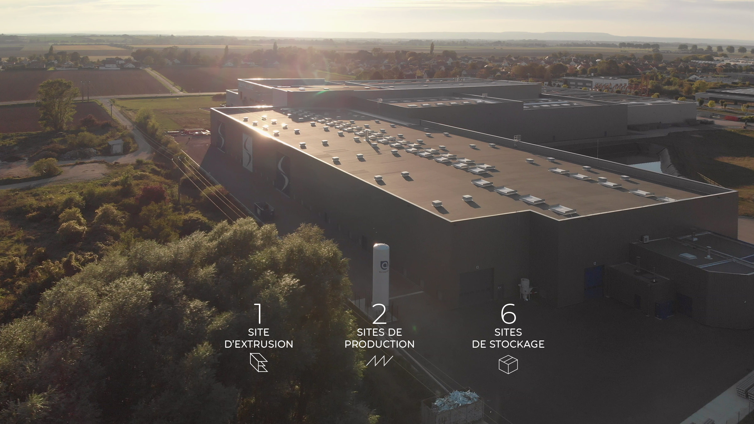 Vue aérienne d'un grand bâtiment industriel entouré de champs, avec des icônes et du texte en français mettant en évidence 1 site d'extrusion, 2 sites de production et 6 sites de stockage - idéal pour une présentation visuelle d'une agence de communication BTP.