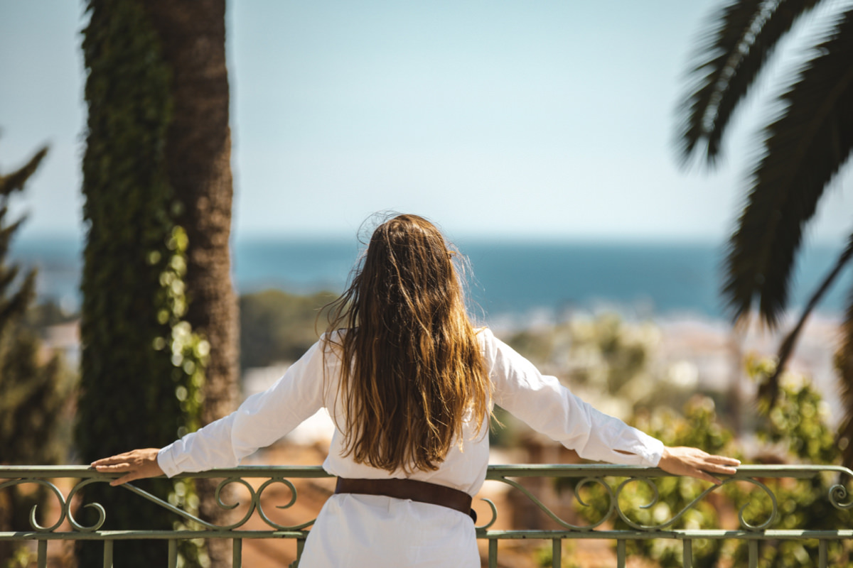 Une femme aux longs cheveux bruns et à la robe blanche se tient sur un balcon, en tenant la balustrade, et regarde une vue floue de l'océan et des feuilles de palmier - une scène inspirante pour la création site internet tourisme lors d'une journée ensoleillée.