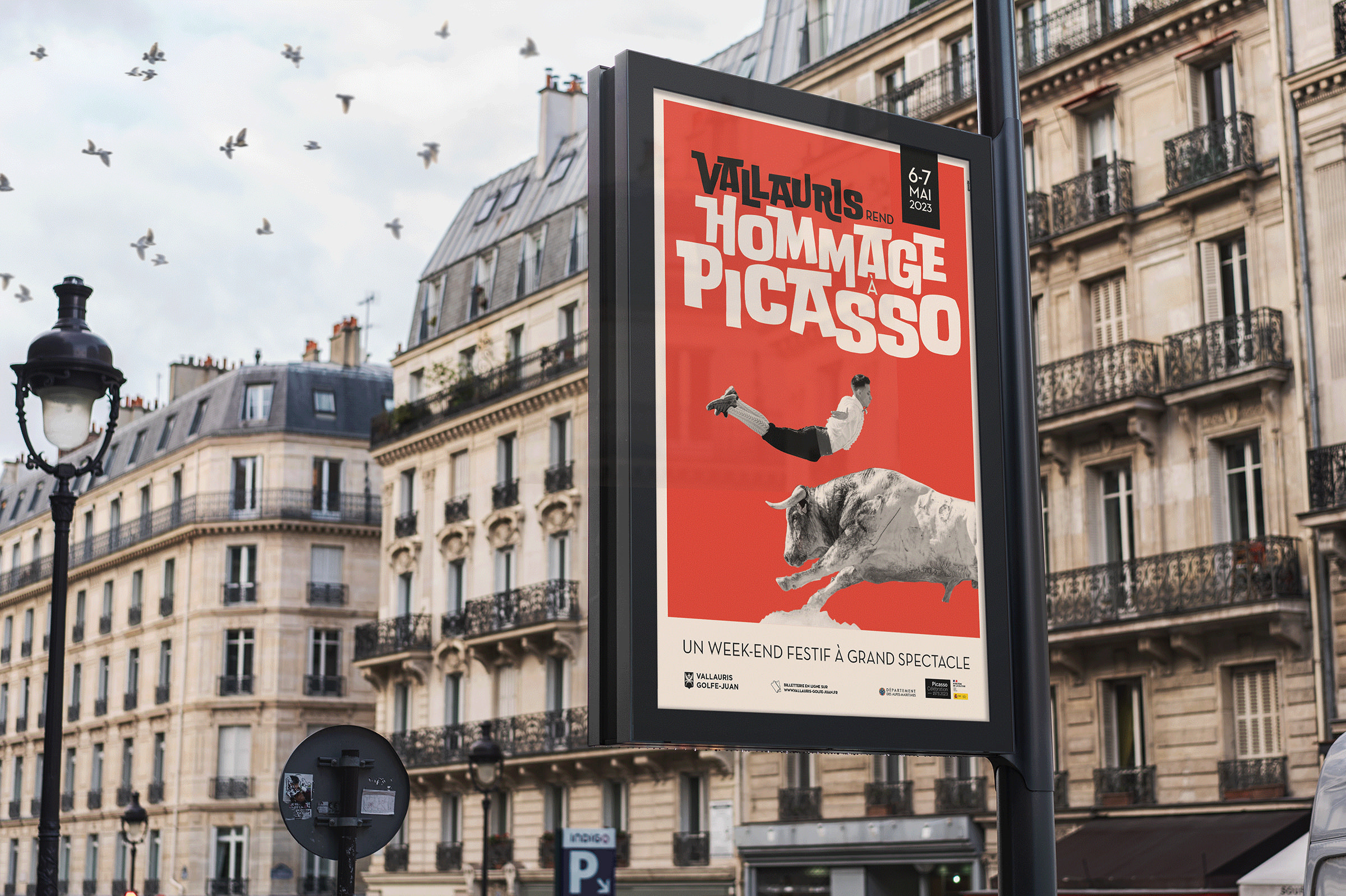 Un panneau d'affichage dans une ville présente une affiche rouge vif pour "Vallauris Hommage Picasso", mêlant art et communication événementielle, avec un homme chevauchant un taureau, des pigeons s'envolant et des bâtiments français classiques en arrière-plan.