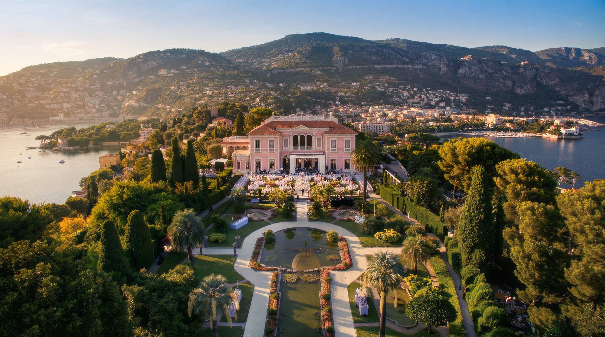 Vue aérienne d'une grande villa entourée de jardins luxuriants, de fontaines et d'allées - un cadre inspirant pour la création d'identité de marque - située sur une colline surplombant une baie, avec des montagnes et une ville en arrière-plan sous un ciel clair.
