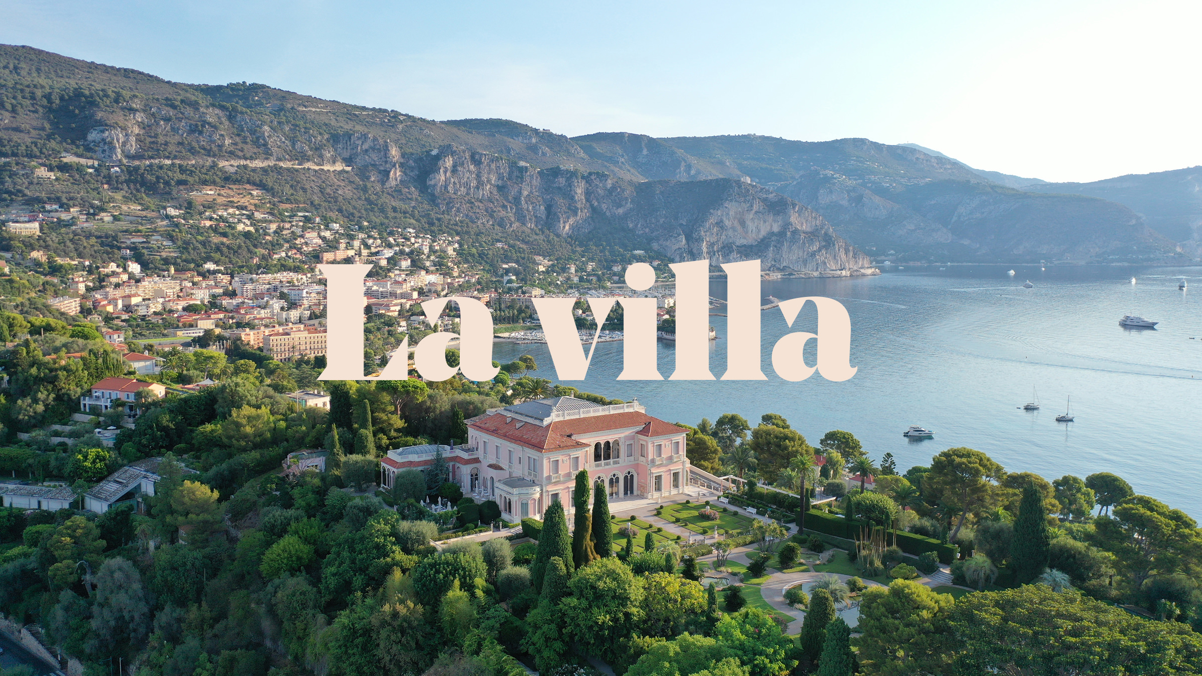 Vue aérienne d'une grande villa entourée d'une végétation luxuriante près de la côte, avec des montagnes et une ville en arrière-plan. Un texte en gros caractères, "La villa", est superposé au centre, ce qui est parfait pour une campagne d'une agence de communication à Cannes.